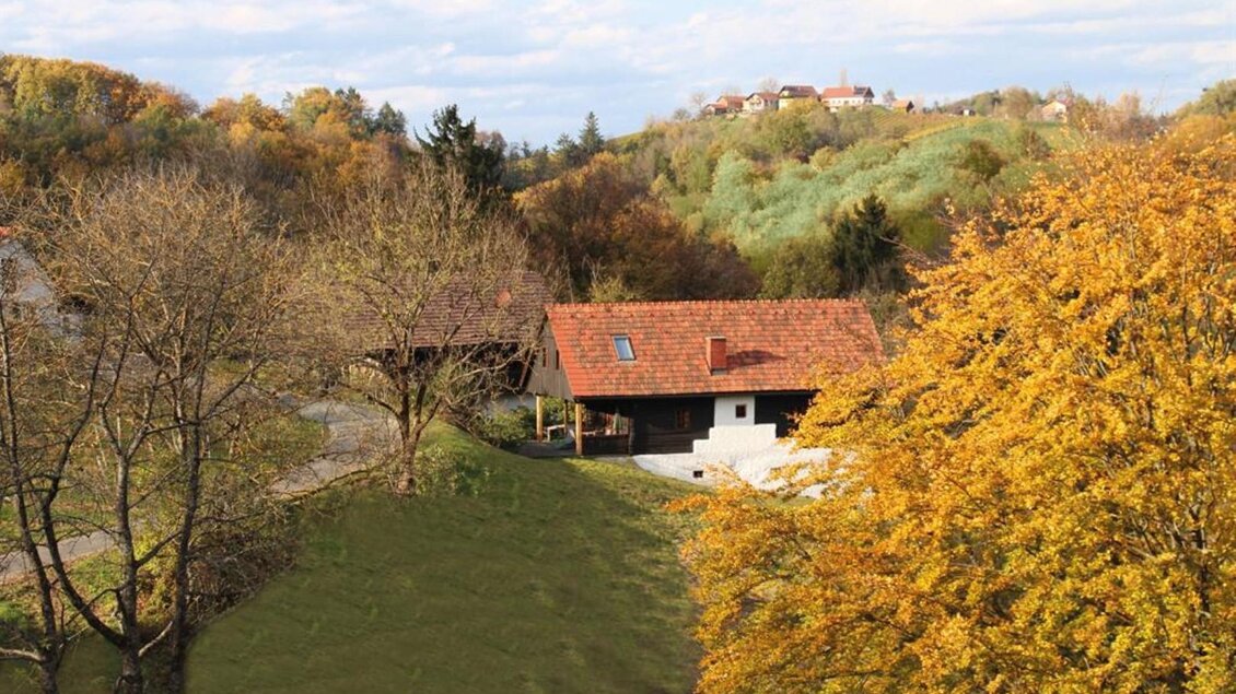 Eine ländliche Szene mit einem kleinen Haus und bunten Herbstbäumen. Im Hintergrund sind sanfte Hügel zu sehen. | © Pongratz