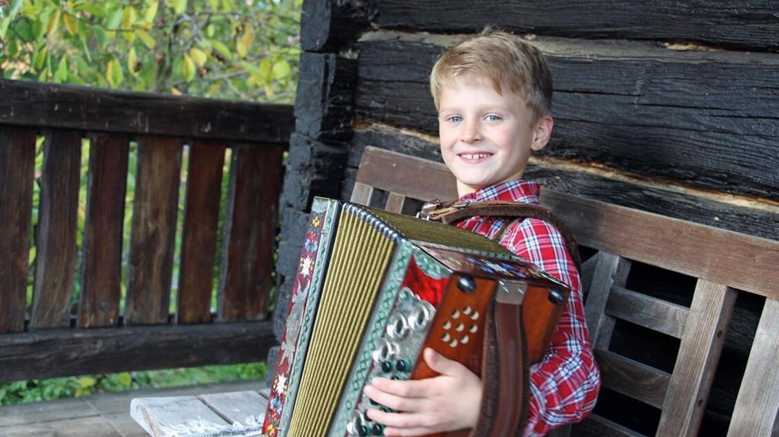 Ein Junge sitzt auf einer Bank und spielt Akkordeon. Er trägt ein kariertes Hemd und lächelt fröhlich. | © Pongratz