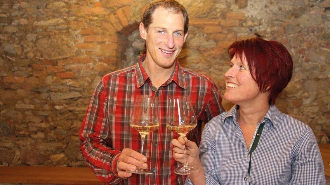 Ein Mann und eine Frau stoßen mit Gläsern an. Sie lächeln sich an und stehen vor einer rustikalen Steinmauer. | © Pongratz