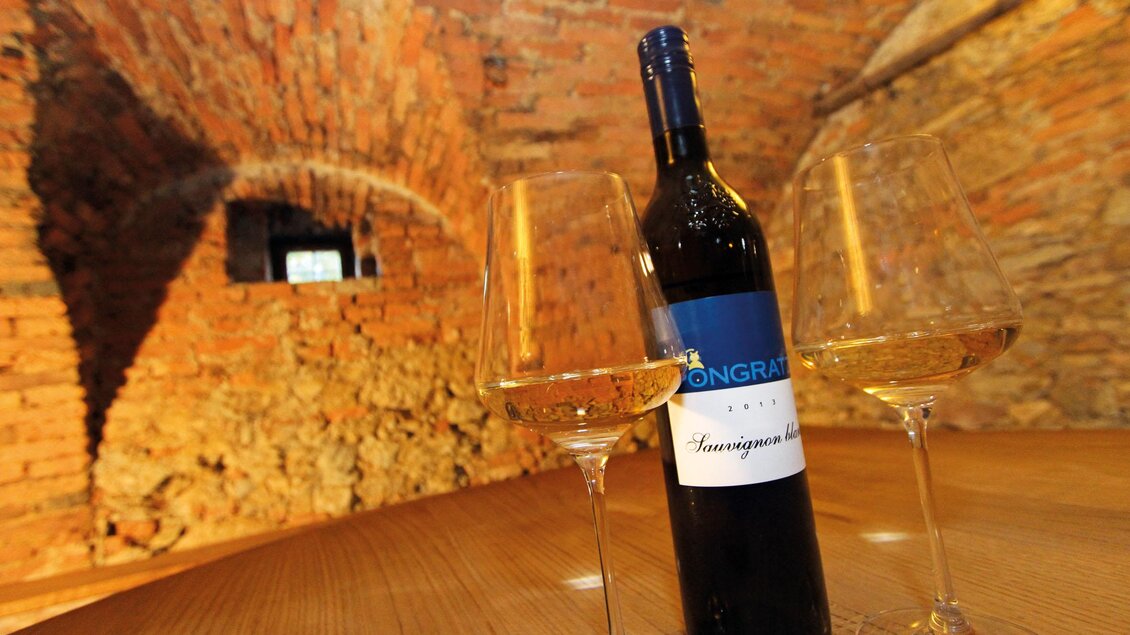 Eine Flasche Sauvignon Blanc steht auf einem Holztisch mit zwei Weingläsern. Der Hintergrund zeigt eine rustikale Kellerwand aus Stein. | © Pongratz