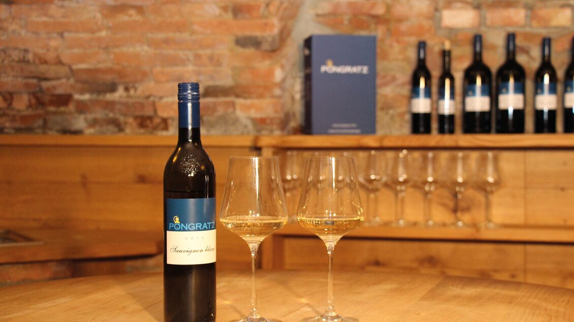 Eine Flasche Wein steht auf einem Holztisch neben zwei Weingläsern. Im Hintergrund sind weitere Weinflaschen und eine elegante Holzvertäfelung zu sehen. | © Pongratz