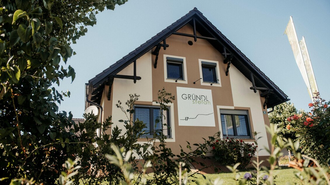 Ein modernes Haus mit einer markanten Fassade und einem Schild, das "GRUNDL" zeigt. Umgeben von grünen Pflanzen und einem klaren Himmel.