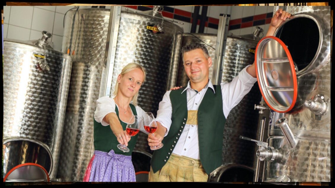 Ein Paar steht in einer Weinkelterei und hält Gläser in der Hand. Im Hintergrund sind silberne Tanks zu sehen. | © Weingut Haubensima