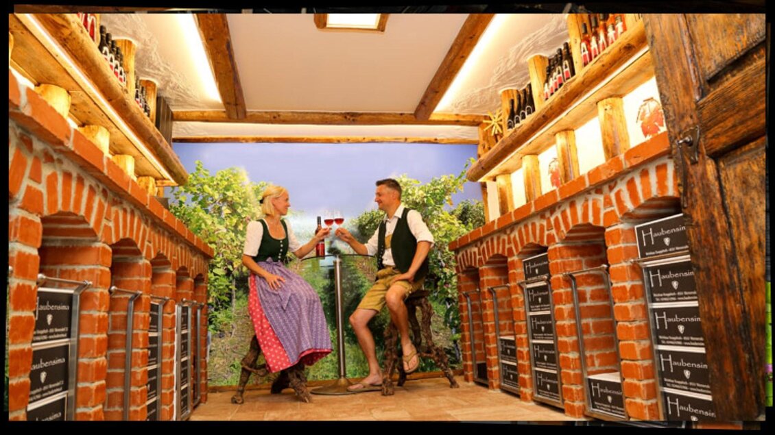Ein gemütlicher Raum mit Ziegelwänden und Regalen voller Flaschen. Zwei Personen in traditioneller Tracht stoßen mit Gläsern an und genießen die Atmosphäre. | © Weingut Haubensima