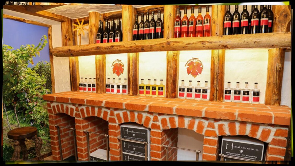 Eine gemütliche Weinbar mit einem Holzregal voller Weinflaschen. Die Wand ist aus Ziegelsteinen und es gibt grünes Pflanzenmaterial im Hintergrund. | © Weingut Haubensima