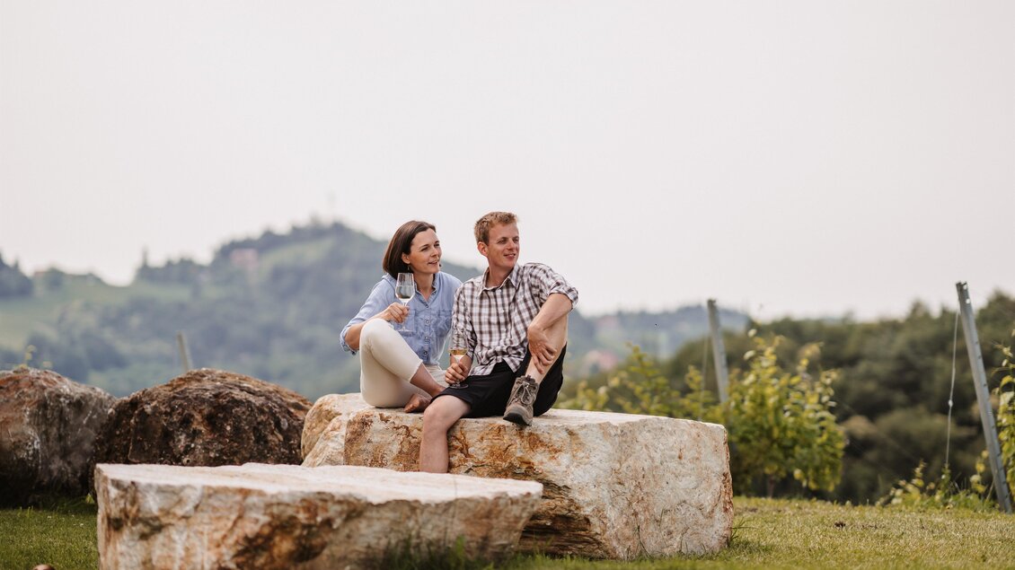 Ein Paar sitzt entspannt auf großen Steinen in einer grünen Landschaft. Im Hintergrund sind sanfte Hügel und Bäume zu sehen. | © MichaelaLorber/WeingutJöbstl