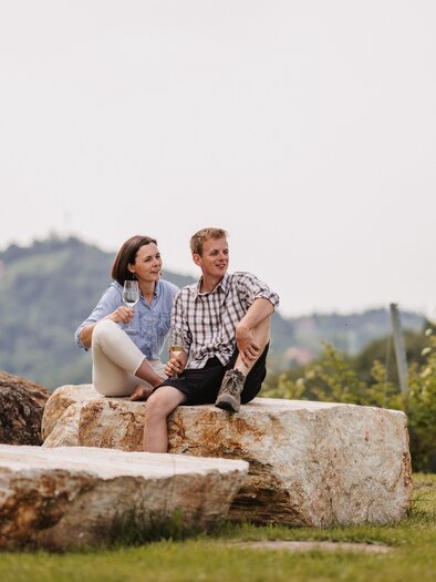 A couple sits relaxed on large stones in a green landscape. In the background, gentle hills and trees can be seen. | © MichaelaLorber/WeingutJöbstl