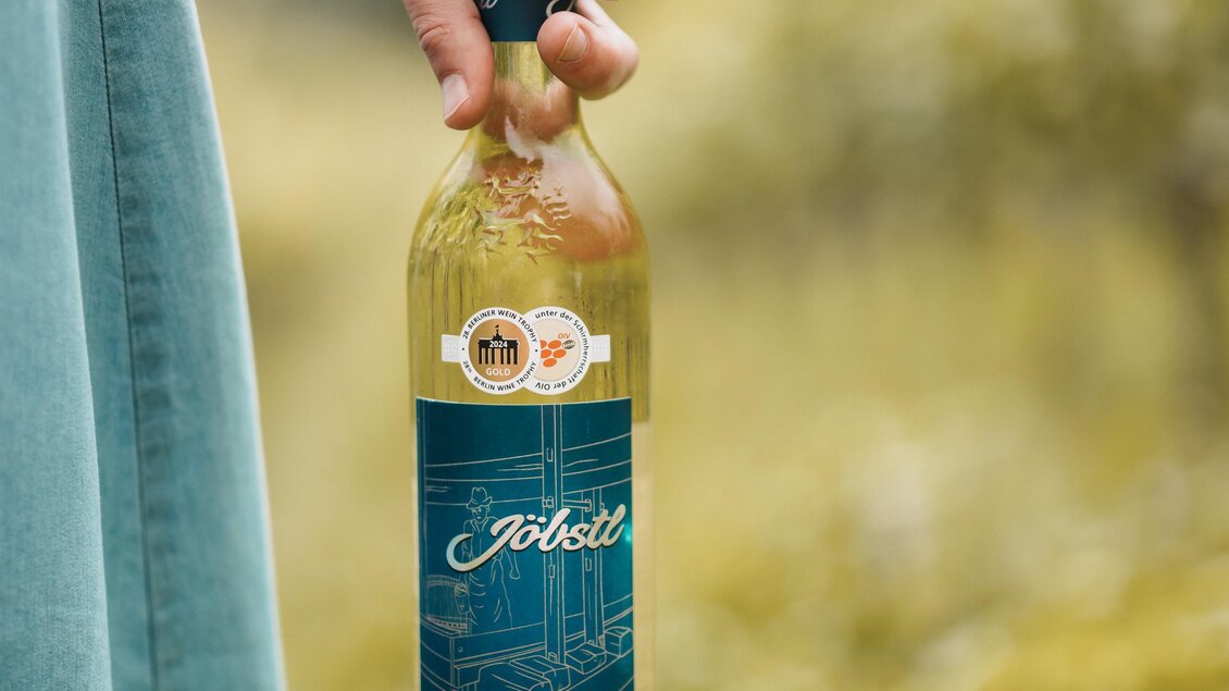 Eine Person hält eine Flasche Weißwein in der Hand. Der Hintergrund zeigt eine grüne, natürliche Landschaft. | © MichaelaLorber/WeingutJöbstl