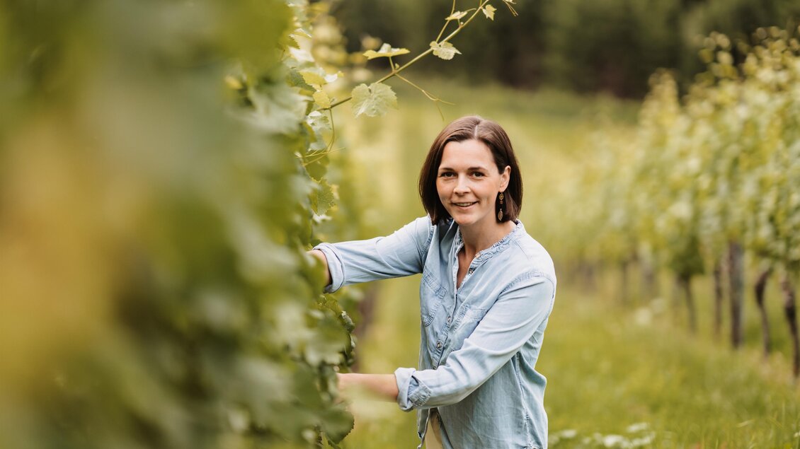 Eine Frau steht in einem Weinberg und pflegt die Weinstöcke. Im Hintergrund sind grüne Reben und eine idyllische Landschaft zu sehen. | © MichaelaLorber/WeingutJöbstl