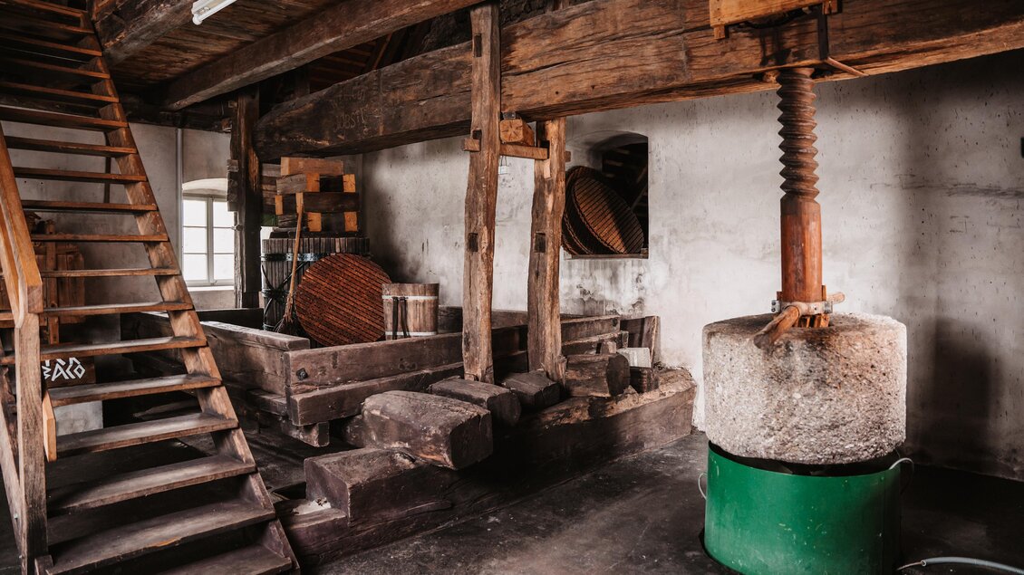 Ein traditionelles Mühlensystem mit einer Holztreppe und einem großen Mühlstein. Die Wände sind aus Stein und die Atmosphäre vermittelt ein historisches Ambiente. | © MichaelaLorber/WeingutJöbstl