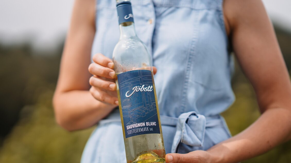 Eine Person hält eine Flasche Sauvignon Blanc in der Hand. Der Hintergrund zeigt eine grüne Landschaft. | © MichaelaLorber/WeingutJöbstl