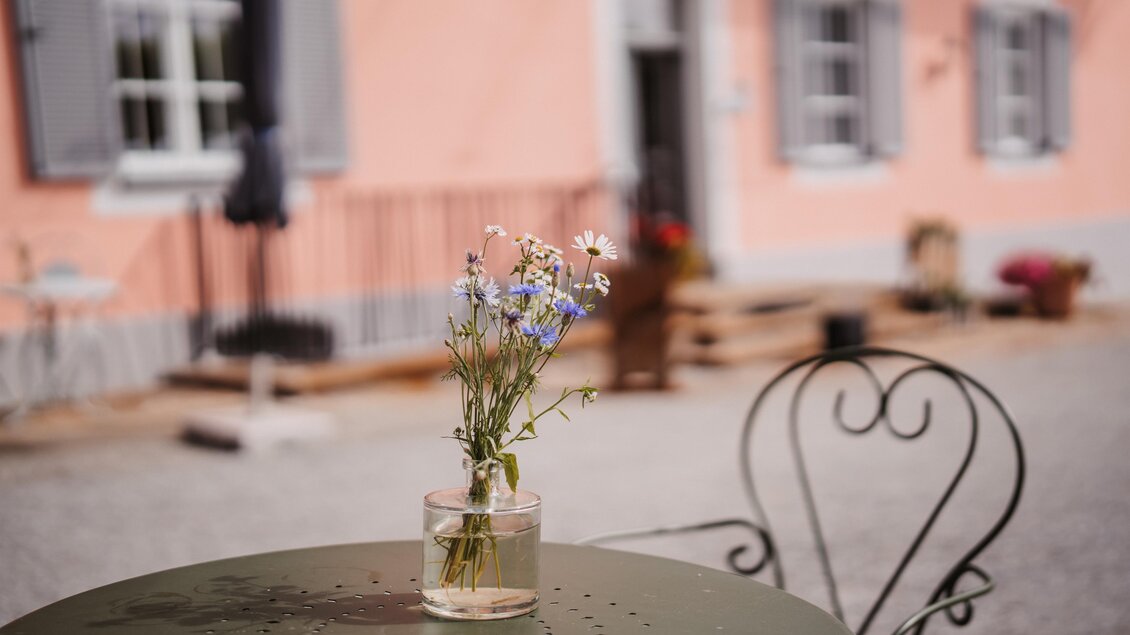 Ein Tisch mit einer Glasvase, die frische Blumen enthält. Im Hintergrund ist ein pinkes Haus mit Fensterläden zu sehen. | © MichaelaLorber/WeingutJöbstl