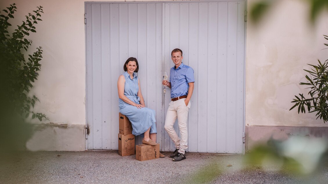 Ein Paar steht vor einer graublauen Tür. Die Frau sitzt auf einem Holzblock, während der Mann neben ihr steht. | © MichaelaLorber/WeingutJöbstl