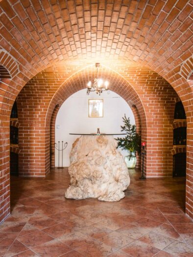 A cozy wine cellar with red brick arches and wooden shelves. In the center stands a large, natural stone, and a green plant adds a touch of nature. | © Riegelnegg