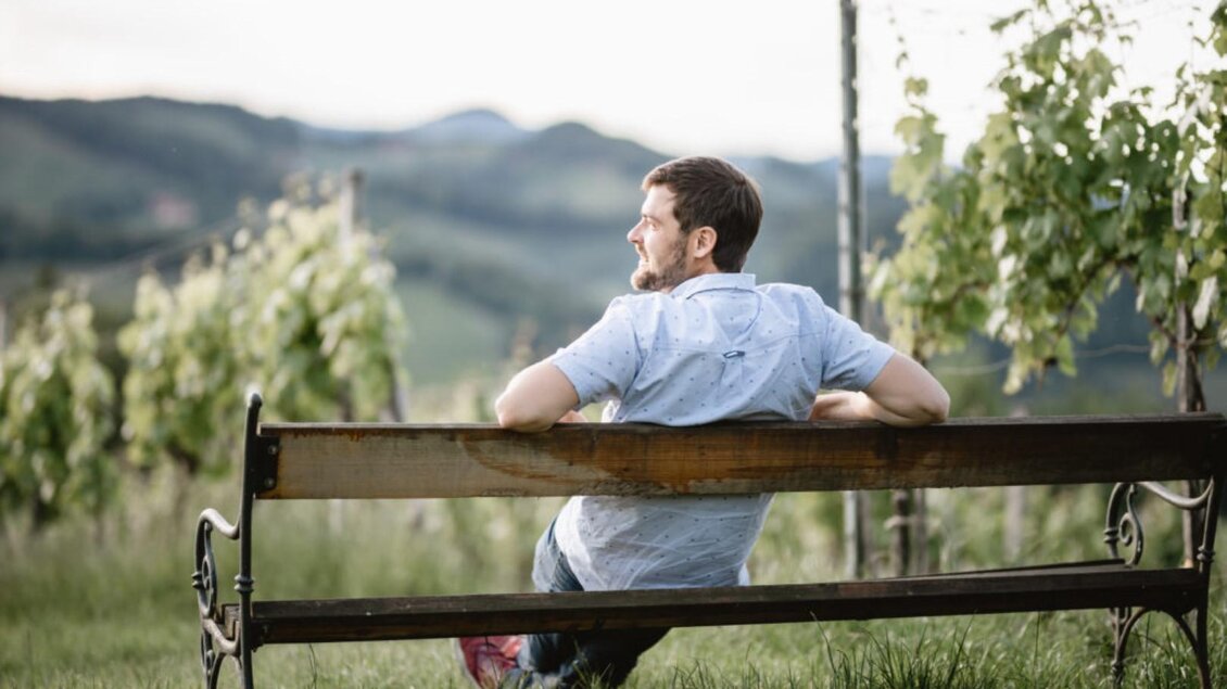 Ein Mann sitzt auf einer Bank in einem Weinberg. Im Hintergrund sind sanfte Hügel und Weinpflanzen zu sehen. | © Riegelnegg