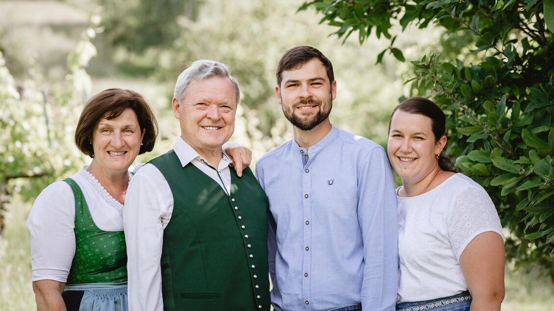 Eine Familie steht lächelnd im Freien vor einer grünen Kulisse. Alle tragen traditionelle Kleidung. | © Riegelnegg