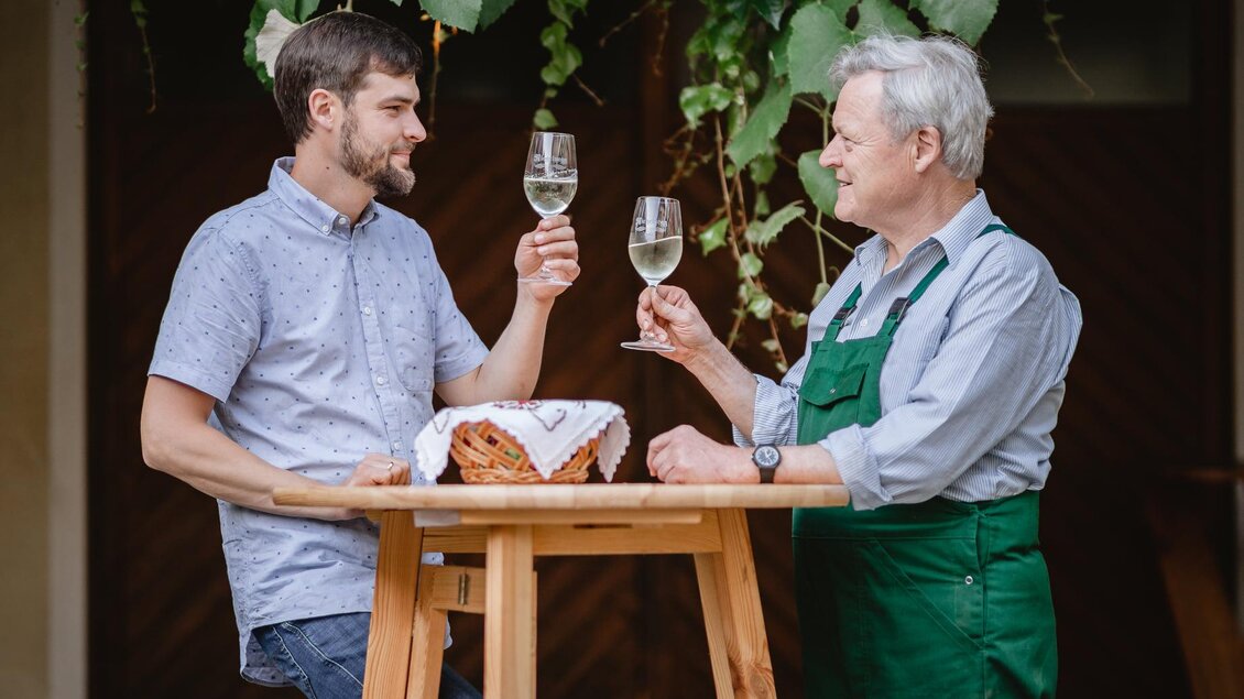 Zwei Männer stoßen mit Gläsern an einem Tisch an. Im Hintergrund sieht man grüne Pflanzen und eine entspannte Atmosphäre. | © Riegelnegg