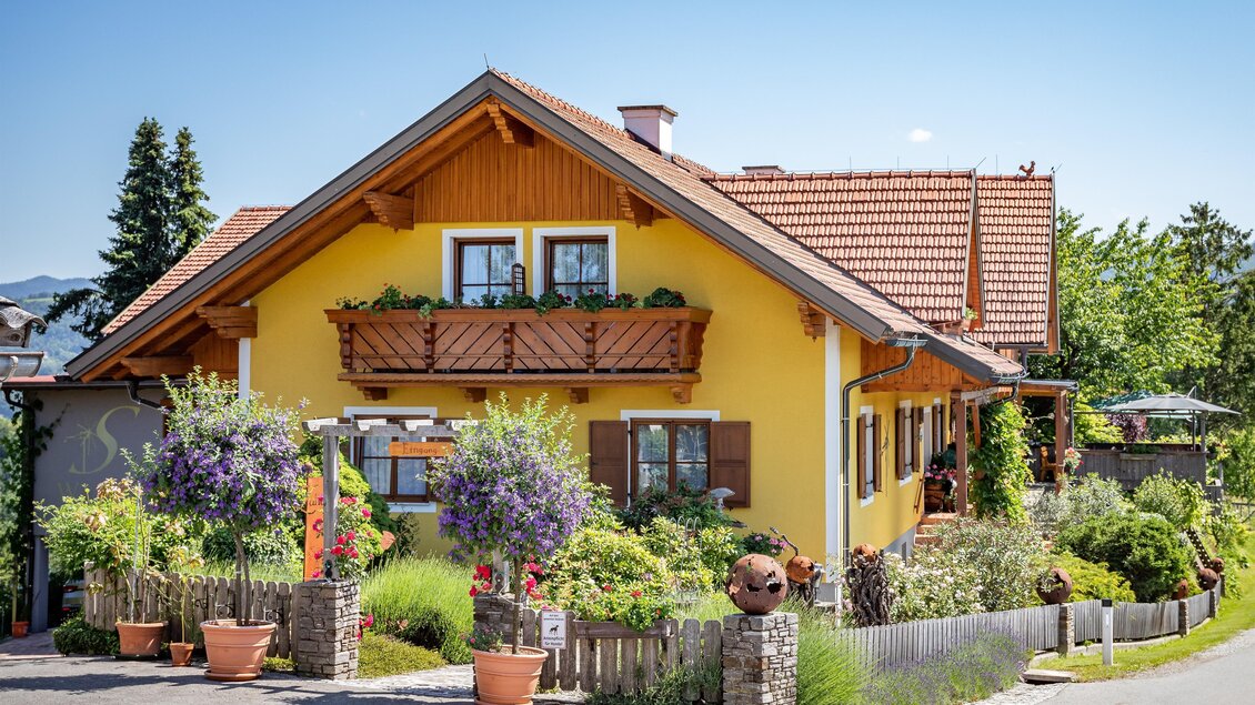 Ein charmantes, gelbes Haus mit einem Balkon und vielen Blumen. Der Garten ist grün und einladend, umgeben von einem Holzzaun. | © Klapotetzhof Silberschneider- Brigitte Maier