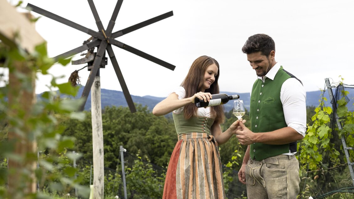 Ein Paar in traditioneller Kleidung steht in einem Weinberg und schenkt Wein in ein Glas ein. Im Hintergrund sind eine Windmühle und sanfte Hügel zu sehen. | © Lisa Resatz_Moonpower