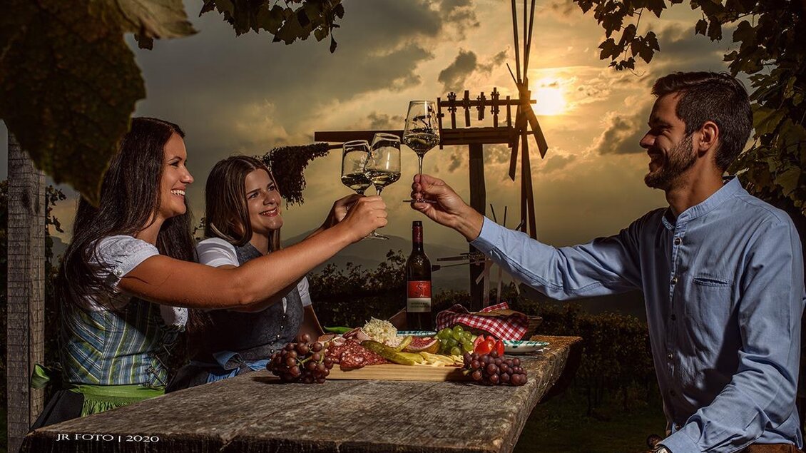 Eine fröhliche Gruppe von drei Personen erhebt Gläser bei Sonnenuntergang. Der Tisch ist mit Wein und frischen Früchten dekoriert, umgeben von Weinreben. | © Jürgen Fuchs | Klapotetzhof Silberschneider