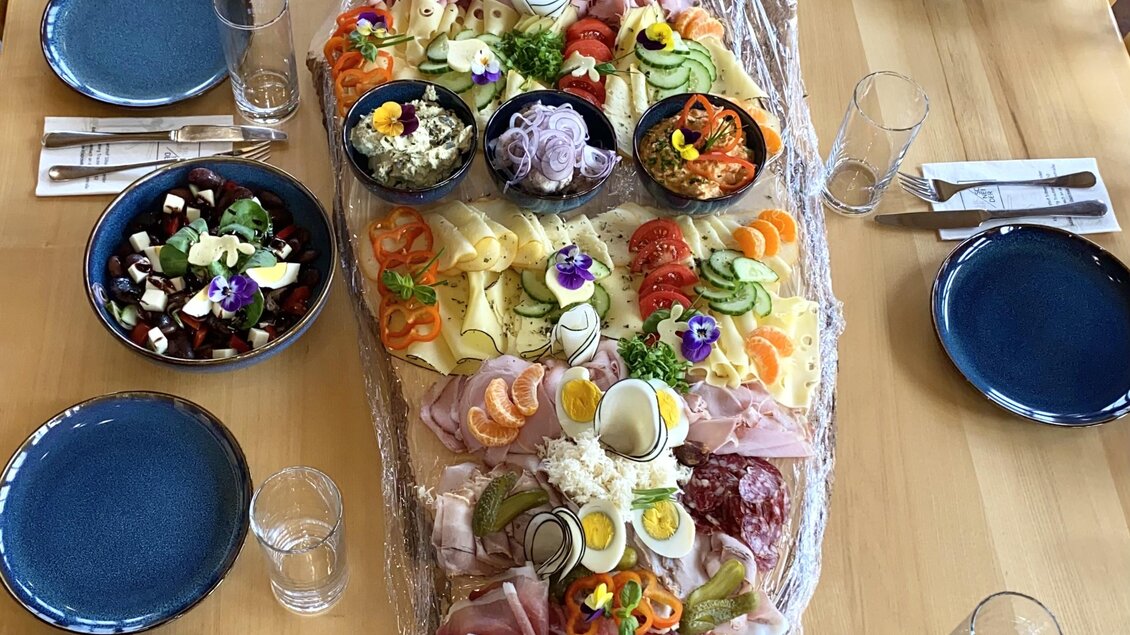 Ein großer Holztisch mit einer langen Platte voller bunter Speisen, einschließlich Wurst, Käse, Gemüse und Salat. Rund um den Tisch stehen Teller mit Salat und Besteck bereit.