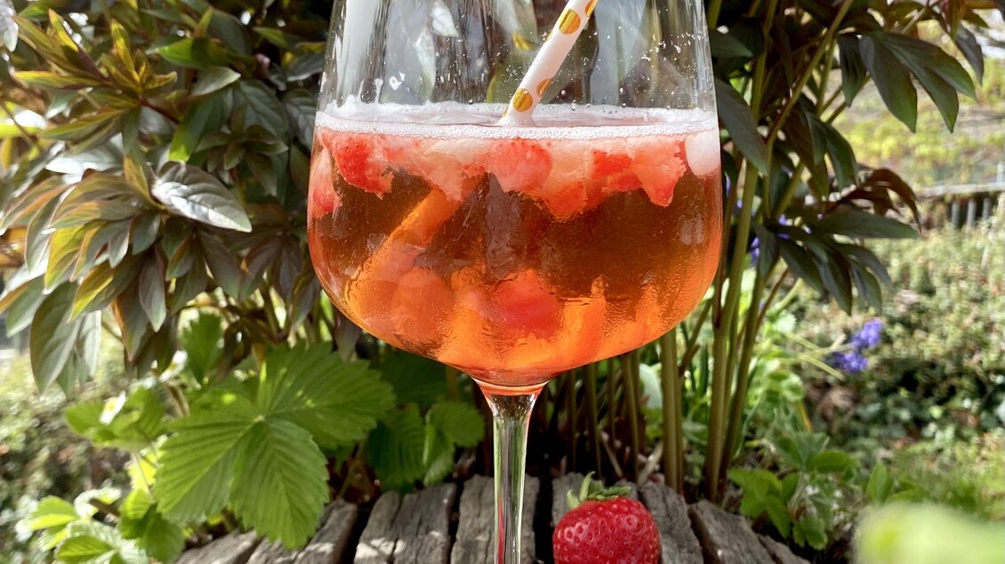 Ein Glas mit einem fruchtigen Getränk und frischen Erdbeeren steht auf einer Holzoberfläche. Im Hintergrund sind grüne Pflanzen zu sehen, was eine entspannte Atmosphäre schafft.