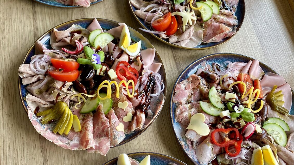 Eine Auswahl an verschiedenen Wurst- und Käseplatten, dekoriert mit frischem Gemüse und Zitrusfrüchten. Die Teller sind ansprechend angerichtet und laden zum Genießen ein.