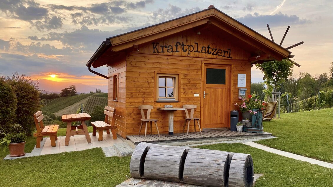 Ein gemütliches Holzhaus mit dem Schriftzug "Kraftplatzerl" steht auf einer Wiese. Im Hintergrund sind sanfte Hügel und ein schöner Sonnenuntergang zu sehen.