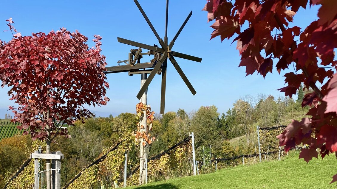 Ein malerischer Garten mit bunten Herbstbäumen und einer alten Windmühle. Die Wiese ist grün und der Himmel ist klar.