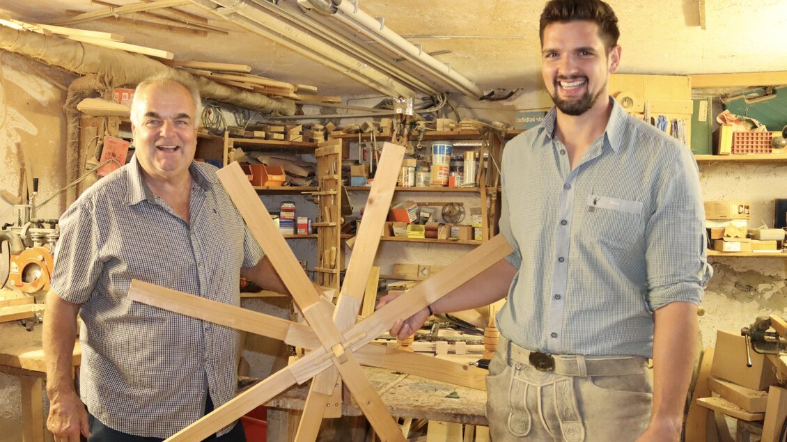 Zwei Männer stehen in einer Werkstatt und halten ein Holzobjekt in Form eines Sterns. Im Hintergrund sind verschiedene Holzwerkzeuge und Materialien sichtbar.