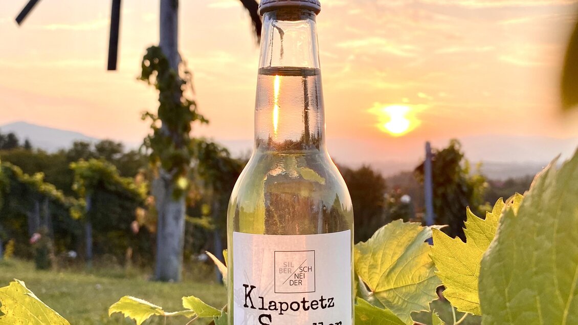 Eine Flasche Klapotetzer Sprudler steht zwischen Weinreben. Im Hintergrund strahlt die Sonne beim Sonnenuntergang.