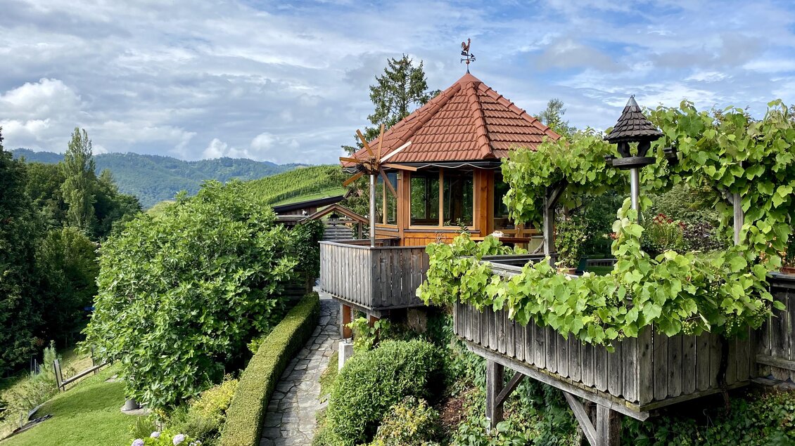Ein gemütliches Holzhaus umgeben von üppigem Grün und Blumen. Im Hintergrund sind sanfte Hügel und ein blauer Himmel zu sehen.