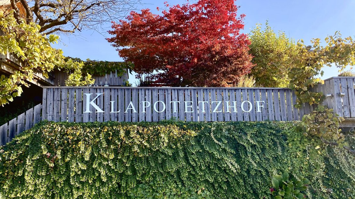 Ein schöner Eingangsbereich mit einem Holzschild, das "Klarenetzhof" zeigt. Im Hintergrund sind grüne Pflanzen und ein roter Baum zu sehen.