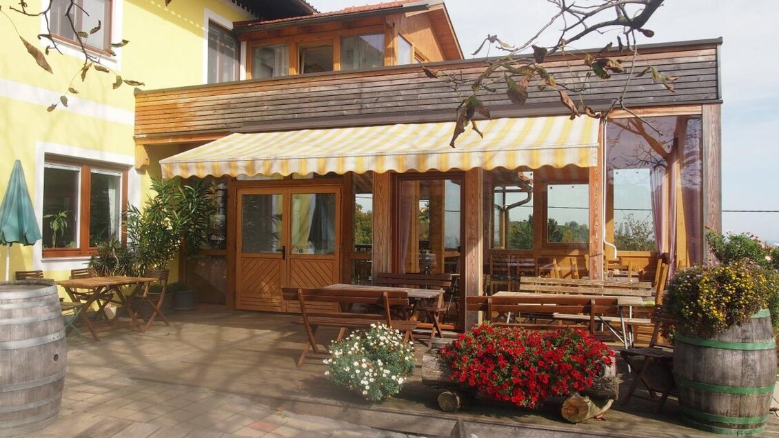 Eine gemütliche Terrasse mit einem Sonnenschirm und rustikalen Holzmöbeln. Bunte Blumen und Pflanzen verleihen dem Außenbereich einen einladenden Charme. | © Weingut Koller