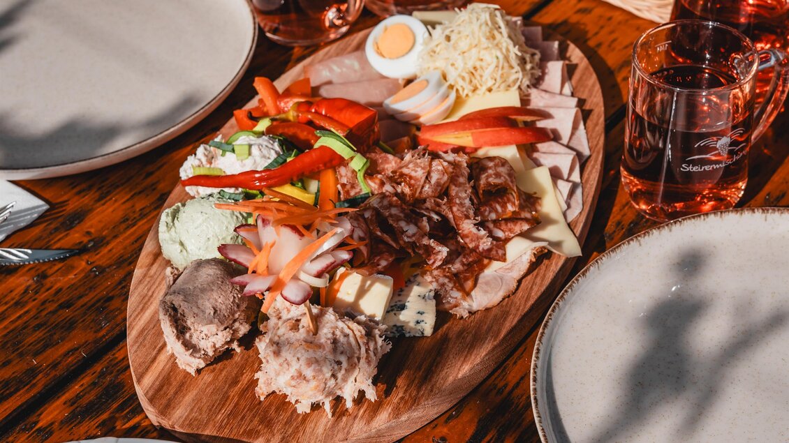 Ein reichhaltiges Essen auf einem Holztisch, bestehend aus verschiedenen kalten Platten mit Fleisch, Gemüse und einem gekochten Ei. Dazu gibt es frisches Brot und Getränke in Gläsern. | © Thermen- & Vulkanland