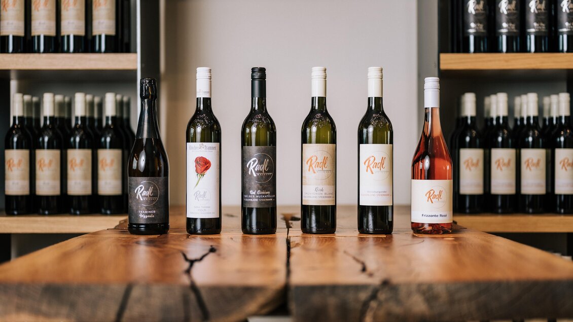 Eine Reihe von sechs Weinflaschen steht auf einem Holztisch. Im Hintergrund sind weitere Weinflaschen auf Regalen sichtbar. | © Anton Meyer - Weingut Radl