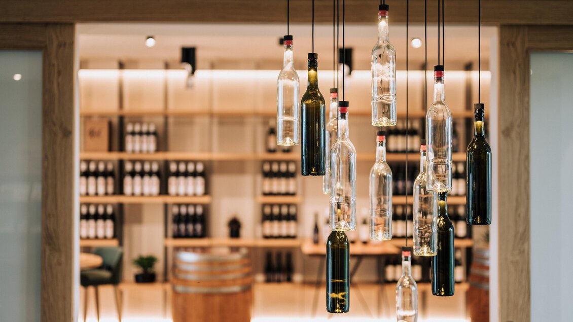 Ein moderner Weinkeller mit hängenden Flaschen aus Glas und einer stilvollen Holzregalwand im Hintergrund. Es gibt auch ein Weinfass, das eine einladende Atmosphäre schafft. | © Anton Meyer-Weingut Radl
