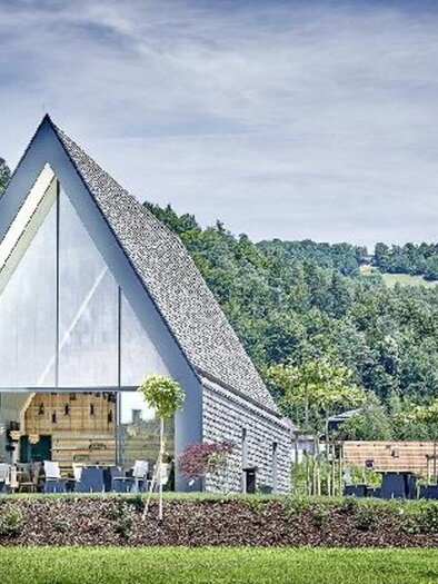 Ein modernes, dreieckiges Gebäude mit großen Fenstern in einer grünen Landschaft. Die harmonische Bauweise fügt sich gut in die Umgebung ein. | © Weingut Wutte