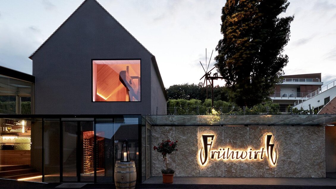 Ein modernes Gebäude mit einer einladenden Fassade. Das Schild „Frühwirt“ ist gut sichtbar und von Pflanzen umgeben. | © Weingut & Buschenschank Frühwirth