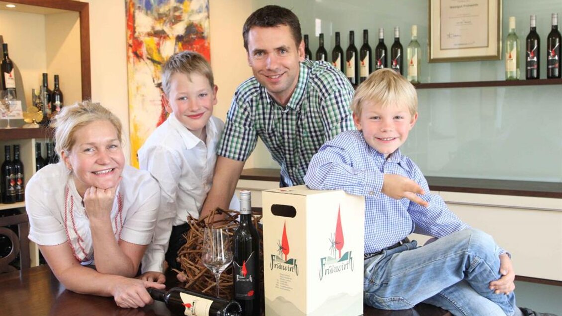 Eine Familie posiert lächelnd in einer Weinbrennerei. Auf dem Tisch stehen Weinflaschen und eine Verpackung. | © Weingut Buschenschank Frühwirt