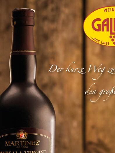 Eine Flasche MARTINEZ Marsala Vergine steht vor einem Holz-Hintergrund. Darunter ist ein Glas und der Slogan "Der kurze Weg zu den großen Weinen" zu sehen. | © Weinhaus der Gallier
