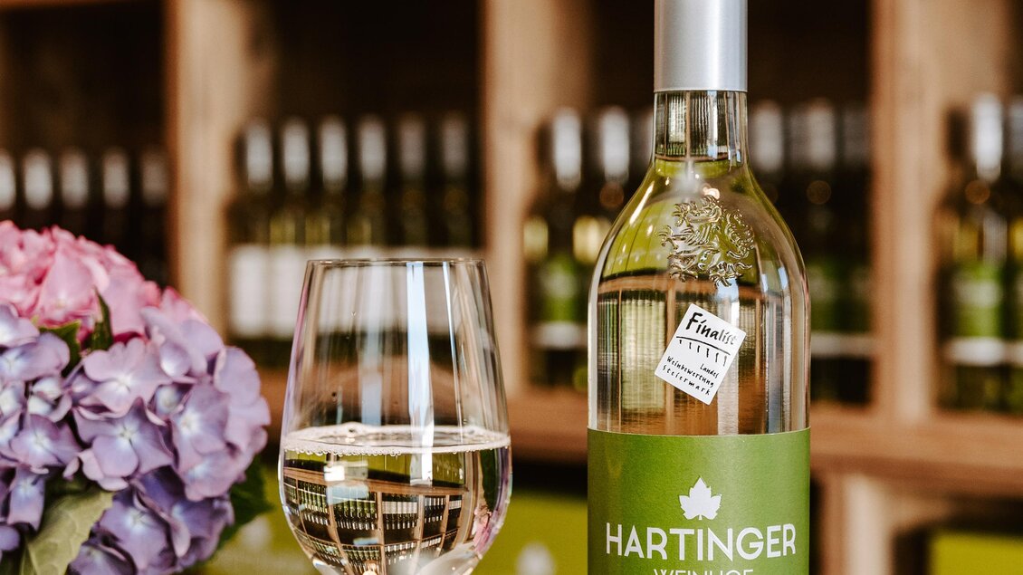 Eine Flasche Hartinger Weißwein steht neben einem Weinglas auf einem Tisch. Im Hintergrund sind Regale mit weiteren Weinflaschen und einer blühenden Pflanze zu sehen. | © Weinhof Hartinger