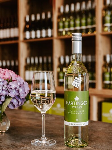 Eine Flasche Hartinger Weißwein steht neben einem Weinglas auf einem Tisch. Im Hintergrund sind Regale mit weiteren Weinflaschen und einer blühenden Pflanze zu sehen. | © Weinhof Hartinger