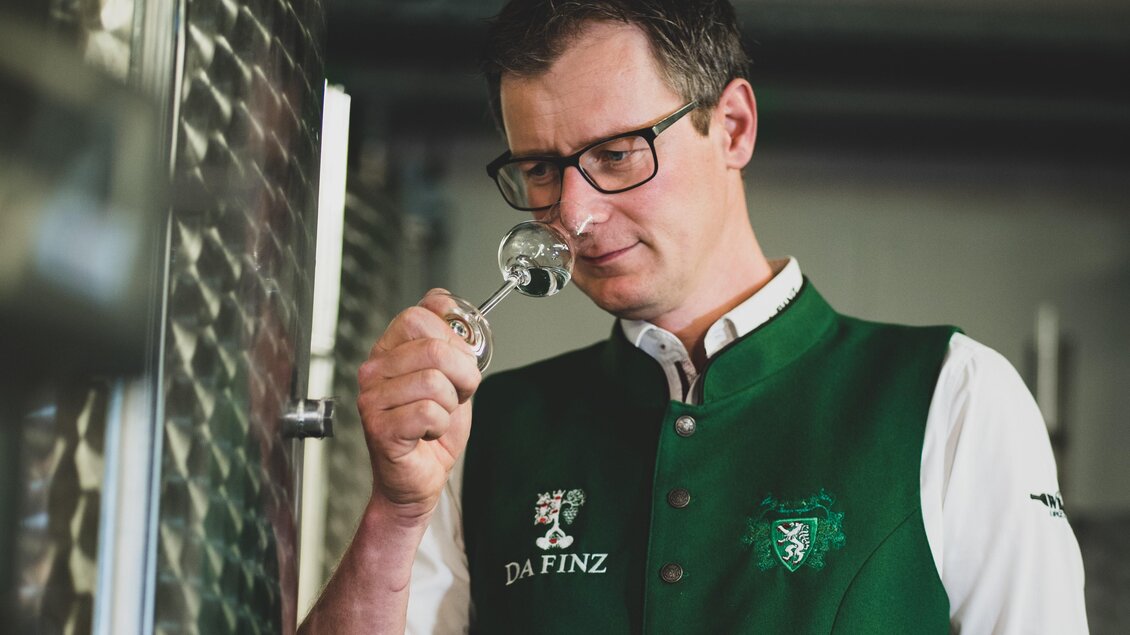 Ein Winzer betrachtet sorgfältig ein Probenröhrchen. Er trägt eine grüne Weste und steht in einem Weinkeller. | © Weinhof Destillerie Da Finz