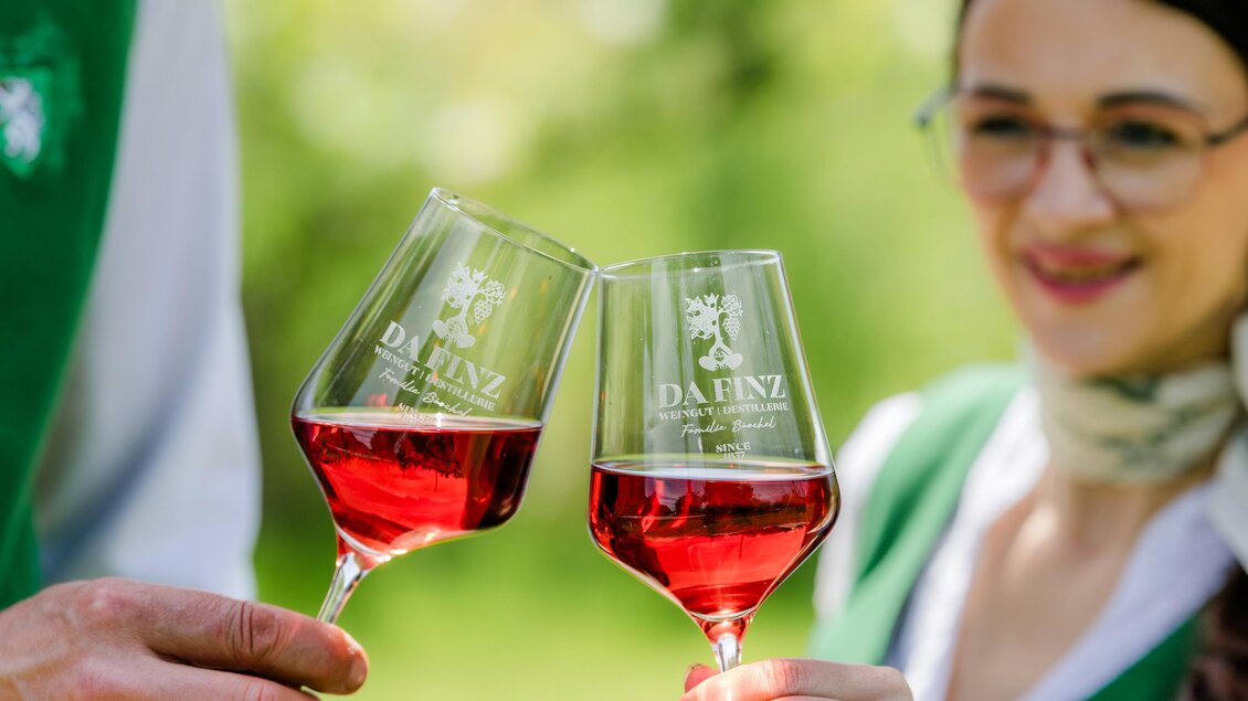 Zwei Weingläser mit roséfarbener Flüssigkeit werden angestoßen. Im Hintergrund sind grüne Wiesen und Bäume zu sehen. | © (c) Region Graz - Mias Photoart