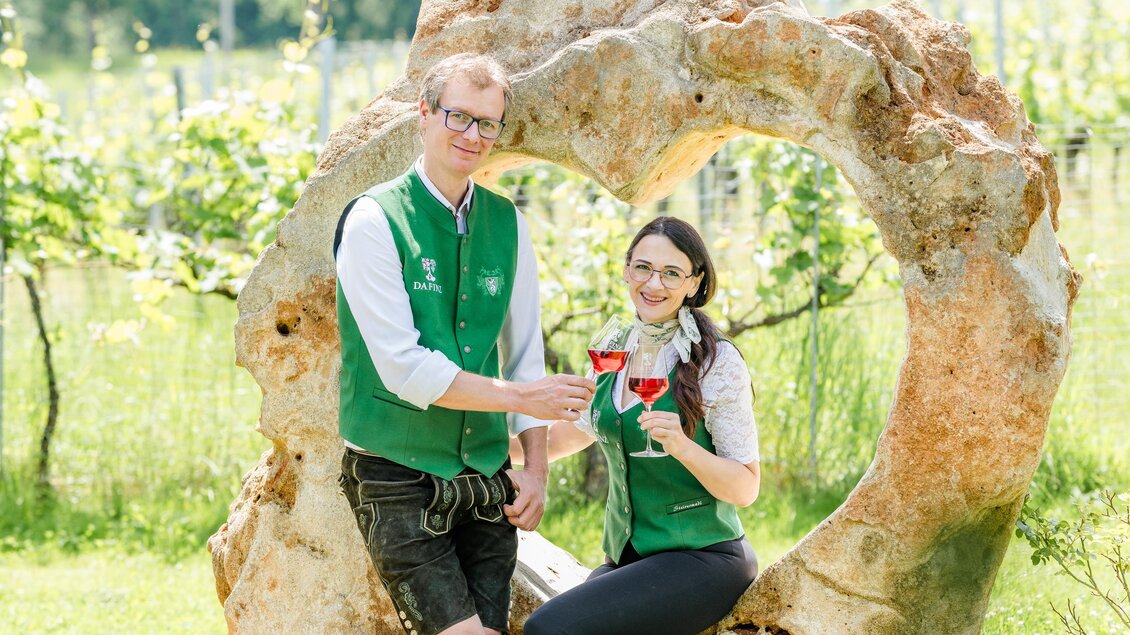 Zwei Menschen heben Gläser in einem Weinberg. Sie stehen vor einem rustikalen Holzrhythmus und strahlen Freude aus. | © (c) Region Graz - Mias Photoart