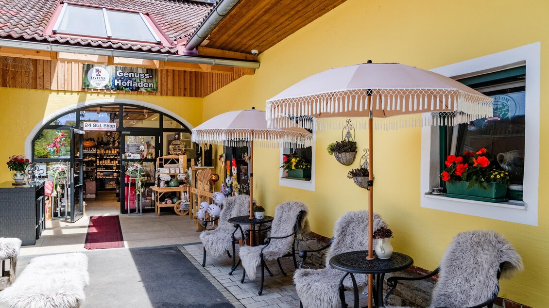 Ein einladender Eingang zu einem Geschäft mit einer gelben Fassade. Vor dem Eingang stehen gemütliche Stühle unter Sonnenschirmen. | © (c) Region Graz - Mias Photoart