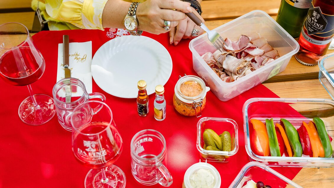 Ein gedeckter Tisch mit einer roten Tischdecke. Es gibt verschiedene Speisen, Getränke und Utensilien, darunter Wurst, Gemüse und Gläser. | © (c) Region Graz - Mias Photoart