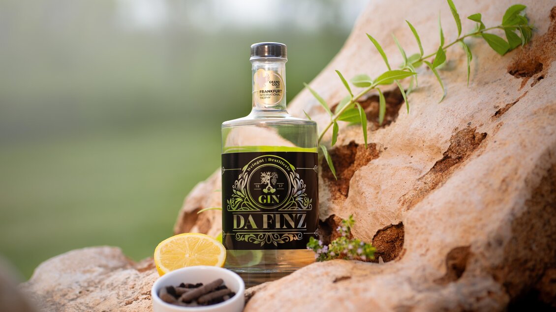 Eine Flasche Gin steht auf einem Holzstück, umgeben von einer Zitrone und Gewürzen. Im Hintergrund ist eine grüne, verschwommene Landschaft zu sehen. | © Weinhof Destillerie Da Finz