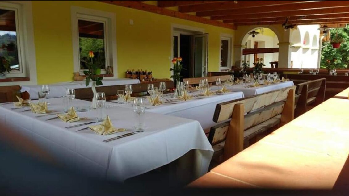 Ein gedeckter Tisch im Freien mit weißen Tischdecken und dekorativen Servietten. Die Umgebung hat gelbe Wände und Pflanzen, die eine einladende Atmosphäre schaffen. | © Buschenschank Heschl